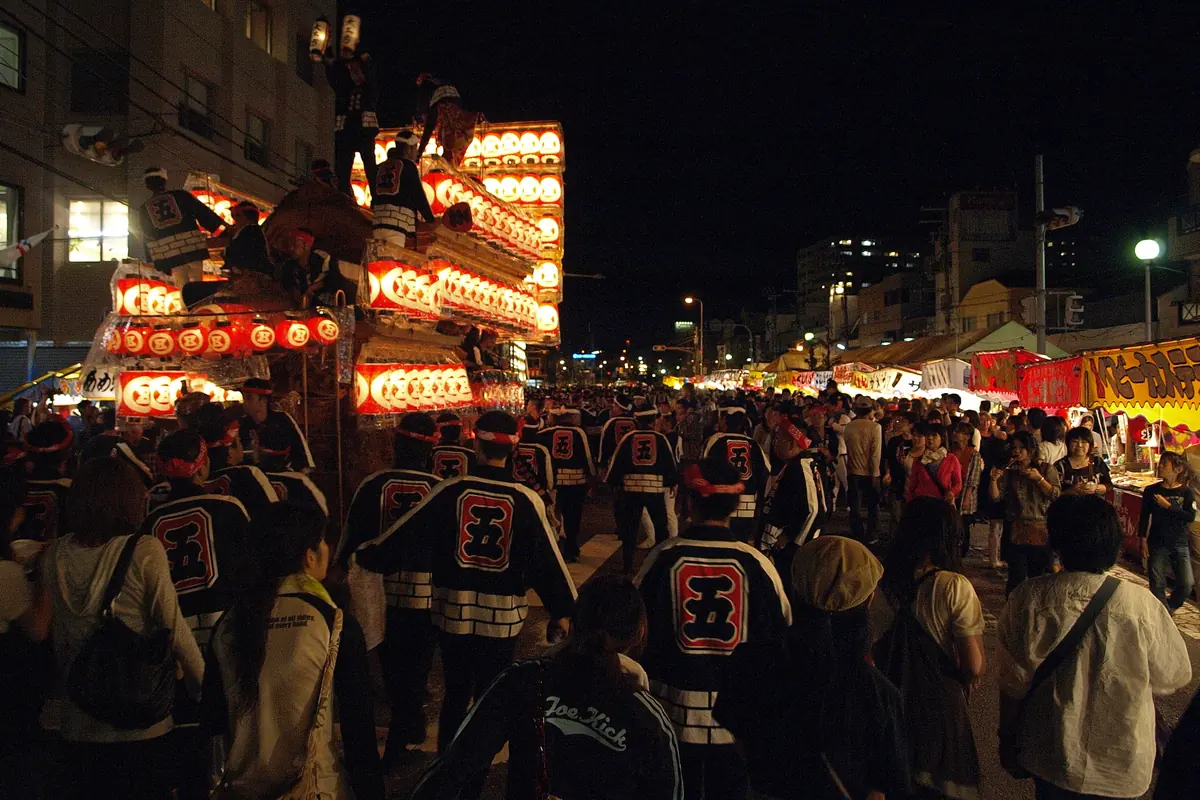 A crowd in happi coats fills a street at night in front of a lit festival float decked with red lanterns