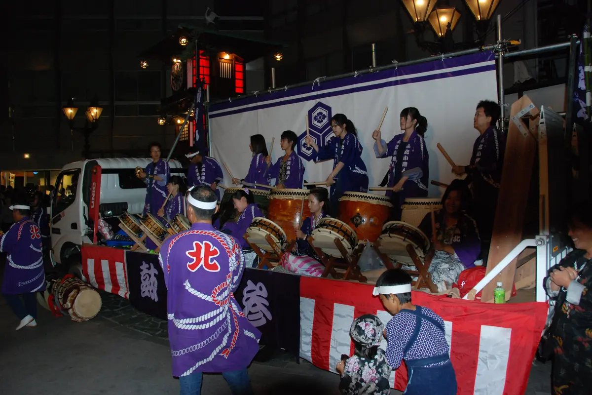 Bubeníci v purpurových festivalových pláštích hrají řadu taiko na ozdobeném voze v noci, pod červeno-bílými závěsy