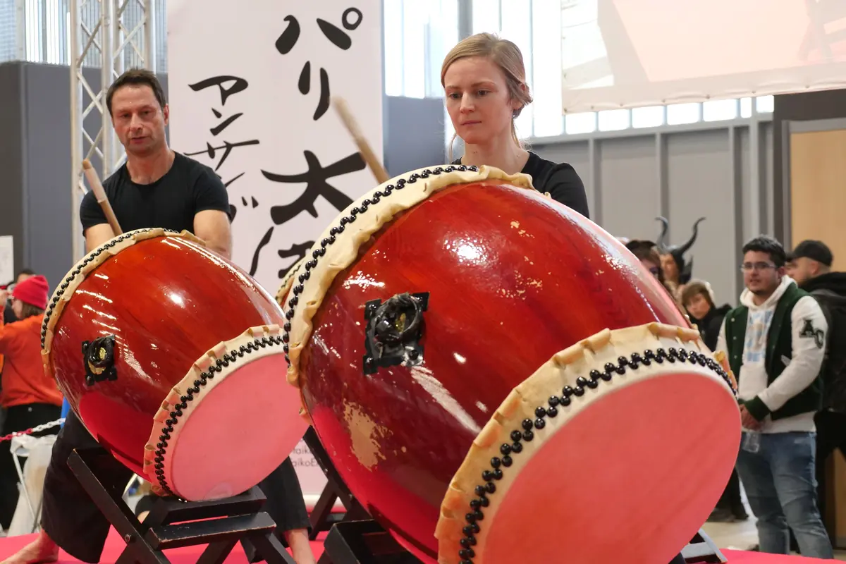 Dva červené taiko bubny s krémovými blánami a řadami cvočků, nakloněné na stojanech, se dvěma bubeníky za nimi