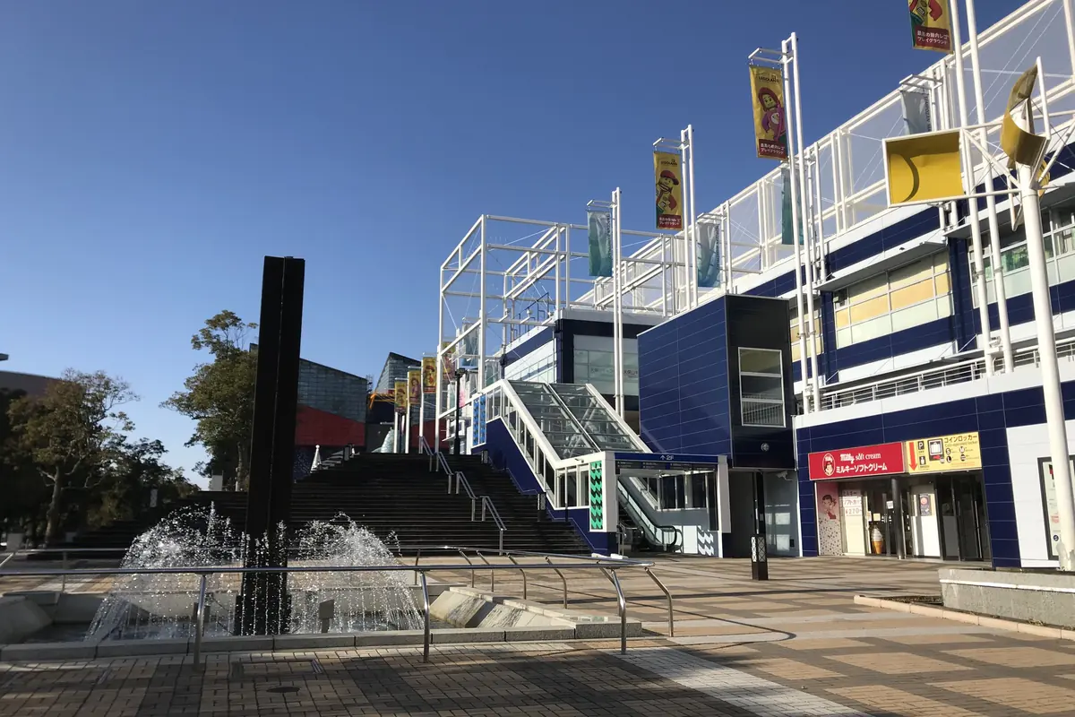 Modrobílá budova Tempozan Marketplace s prapory na vysokých stožárech, vedle fontány na dlážděném náměstí pod jasným nebem